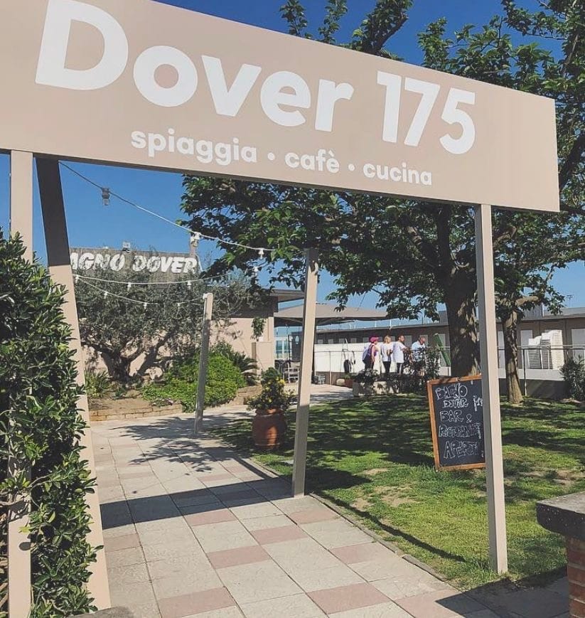 Ingresso del Bagno Dover 175 con la grande insegna 'Dover 175 — spiaggia · cafè · cucina' sospesa sopra il viale fra olivi e siepi